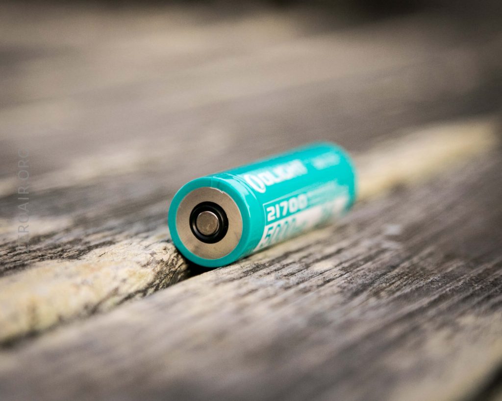 A teal and silver cylindrical 21700 lithium-ion battery lying horizontally on a weathered wooden surface, with the positive terminal facing the camera.