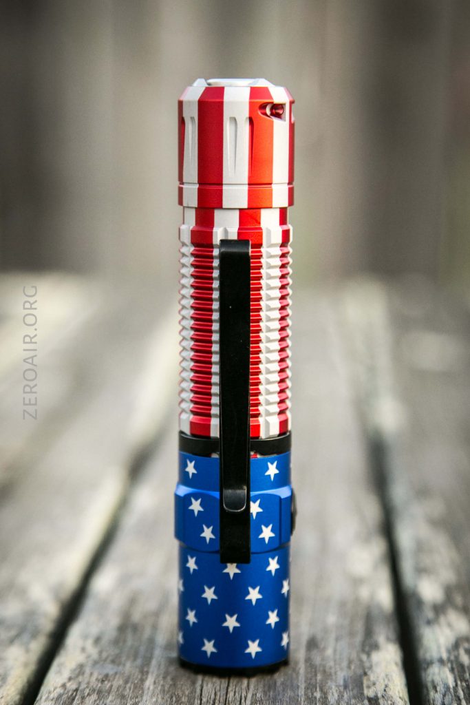 A flashlight standing upright on a wooden surface, featuring a design with red and white stripes on the top and blue with white stars on the bottom, resembling the American flag.