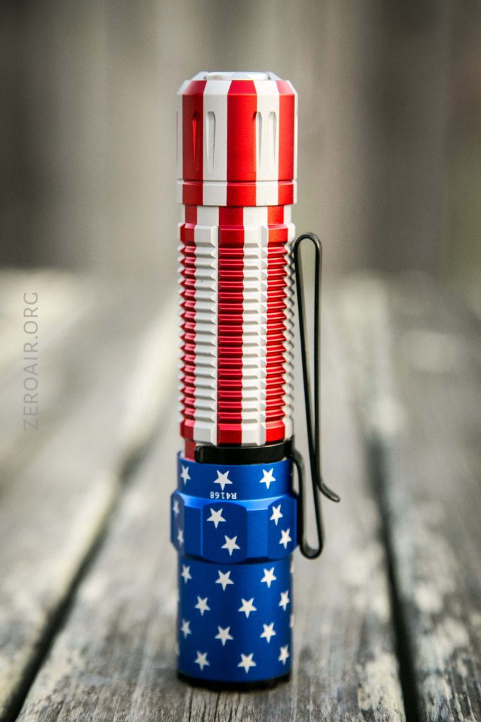 A flashlight with a red, white, and blue design resembling the American flag, standing upright on a wooden surface. The body has red and silver stripes, and the base is blue with white stars.