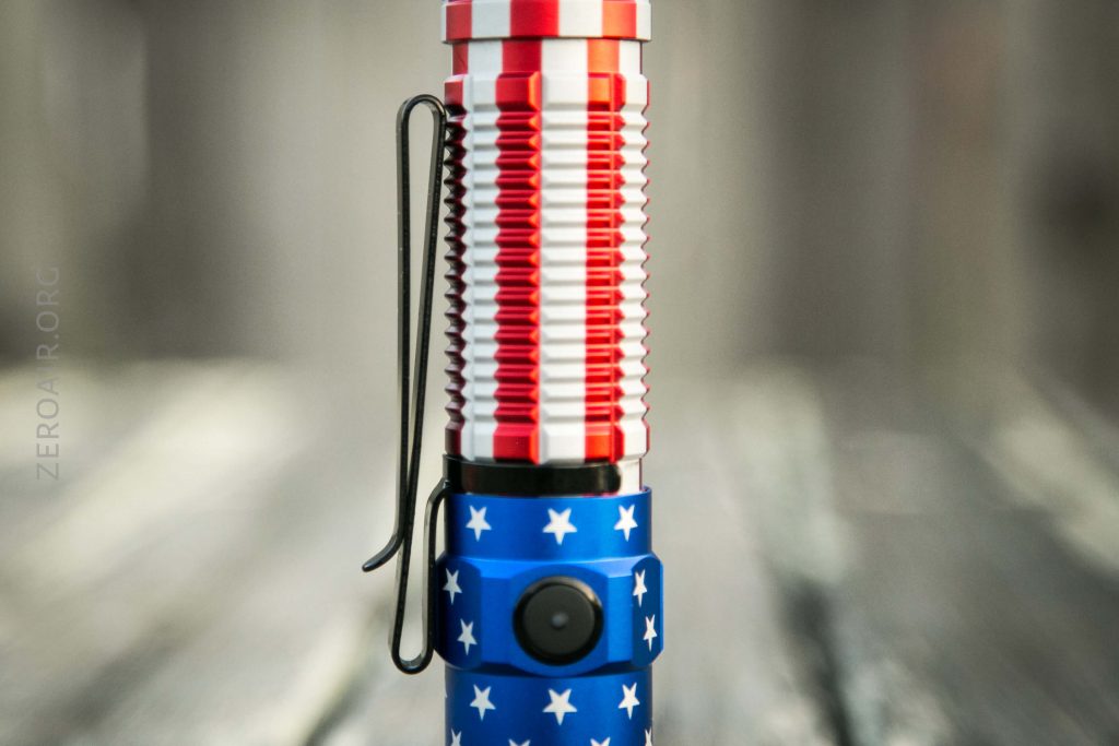 A close-up image of a flashlight with a red, white, and blue design featuring white stars and stripes. The flashlight has a black pocket clip and a button on the blue section.