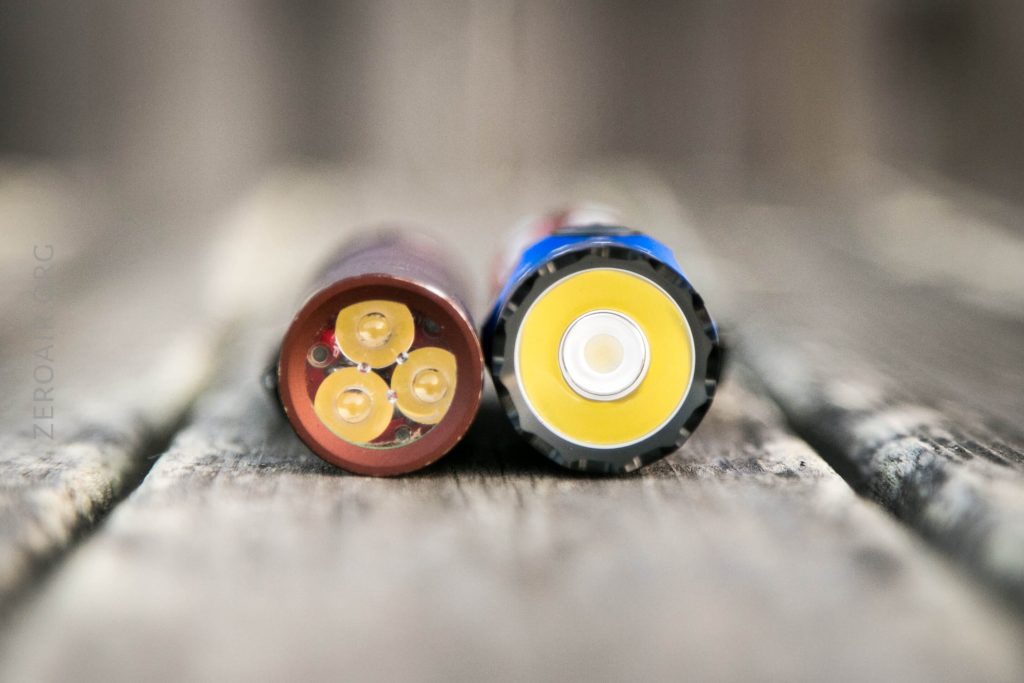 Close-up of the ends of two cylindrical batteries placed side by side on a wooden surface, showing their contact points and different designs.