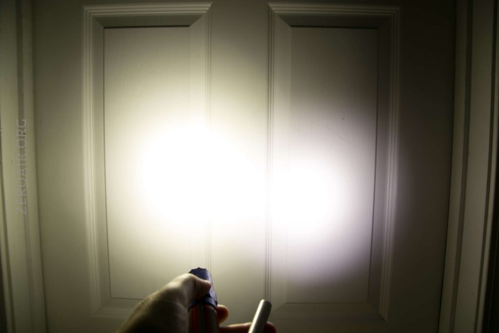 A hand holds a flashlight shining a bright beam of light onto a white, paneled door, creating a strong glare in the center of the image.