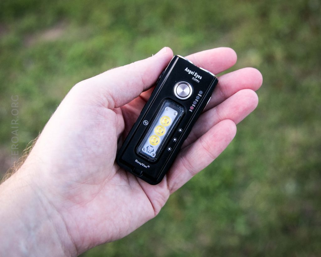 A person holds a small black handheld flashlight called “Angel Eyes” with control buttons and visible LED bulbs, against a blurred grassy background.