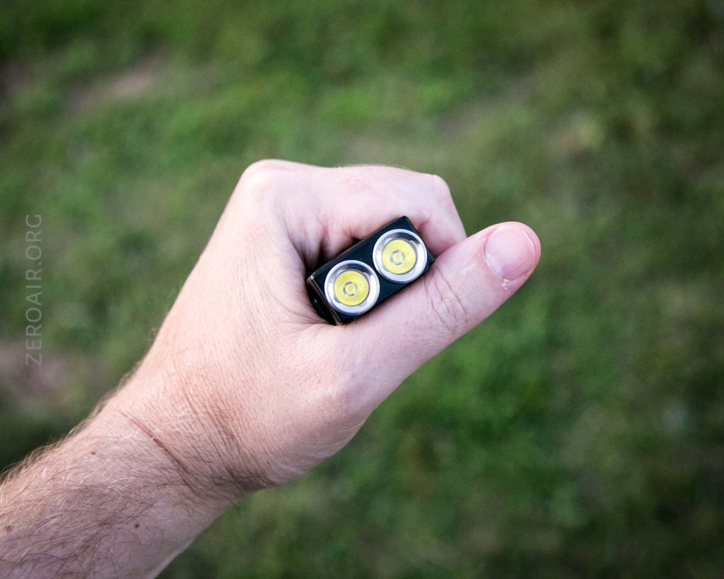 A hand holding a small black flashlight with two yellow LED bulbs, shown from above with a grassy area in the background.