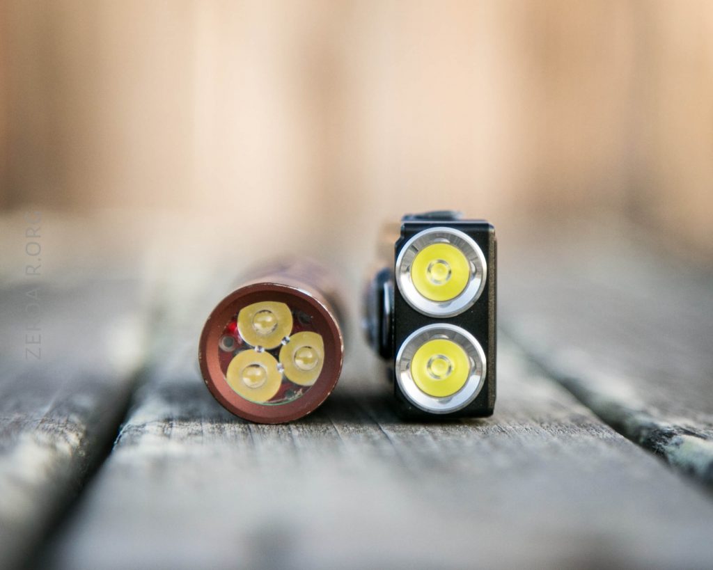 A close-up of two small LED flashlight heads placed side by side on a wooden surface, with a blurred background. The left flashlight is cylindrical with three LED bulbs, and the right one is rectangular with two LED bulbs.