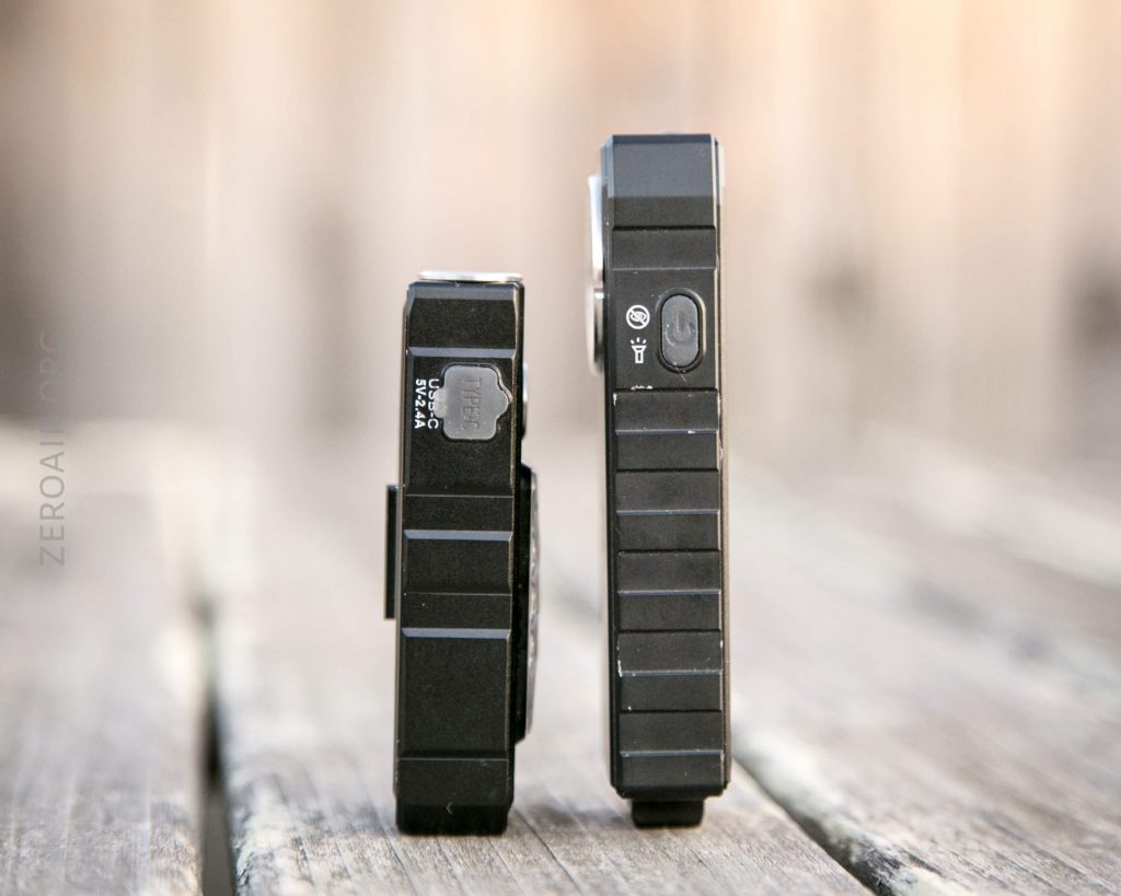 Two black electronic devices stand upright on a wooden surface, viewed from the side. The left device has a clip and a visible SIM card tray; the right device has a SIM icon and appears slightly larger.