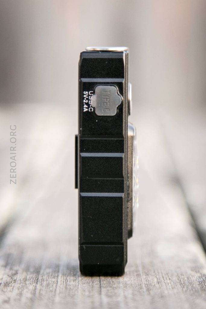 A black electronic device, shown from the side, rests on a wooden surface. The Micro USB port and text USB-C 5V=2A are visible. The background is softly blurred.