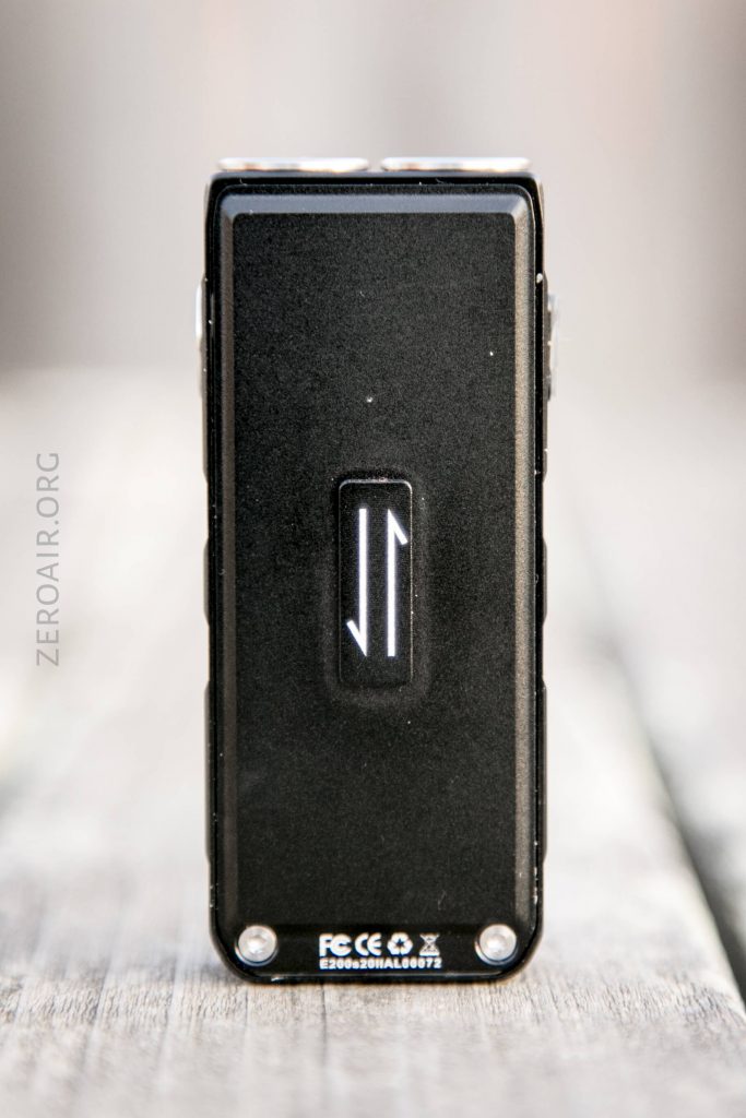 A rectangular black electronic device stands upright on a wooden surface. The front displays a white arrow-like symbol, with certification markings and screws visible at the bottom. The side shows ZEROAIR.ORG faintly.