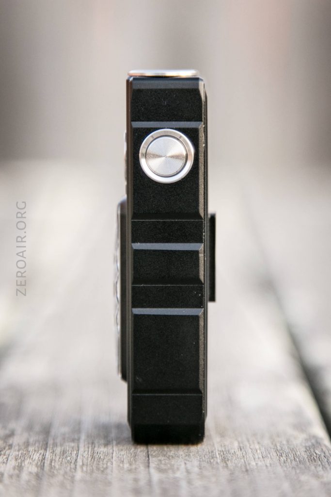 A close-up side view of a black rectangular electronic device with a metallic circular button, resting upright on a wooden surface. The background is blurred and the text zeroair.org appears vertically.