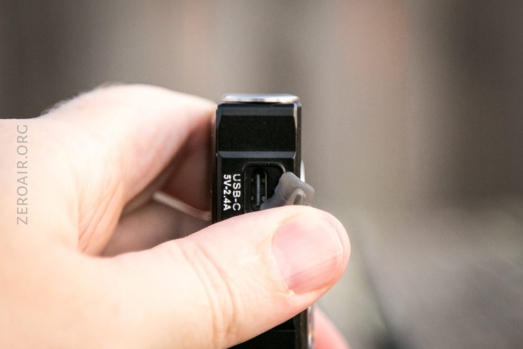 A close-up of a person holding a black electronic device, with a thumb lifting a rubber flap to reveal a USB-C charging port labeled USB-C 5V 2.4A. The background is blurred.