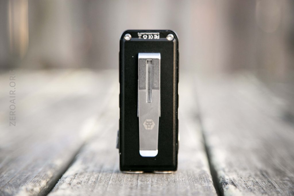 A close-up of a rectangular black device with a metal clip standing vertically on a weathered wooden surface, with a blurred background. The device has visible branding and certification markings on the top edge.