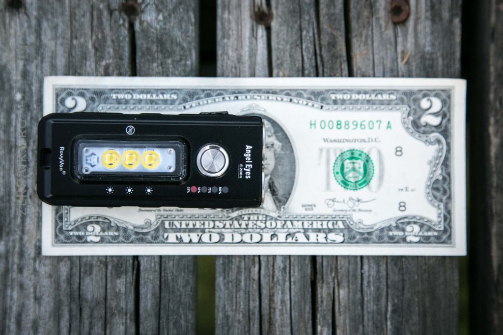 A black Angle Eyes flashlight is placed on top of a two-dollar bill, which lies on a weathered wooden surface. The flashlight and bill are arranged horizontally for size comparison.