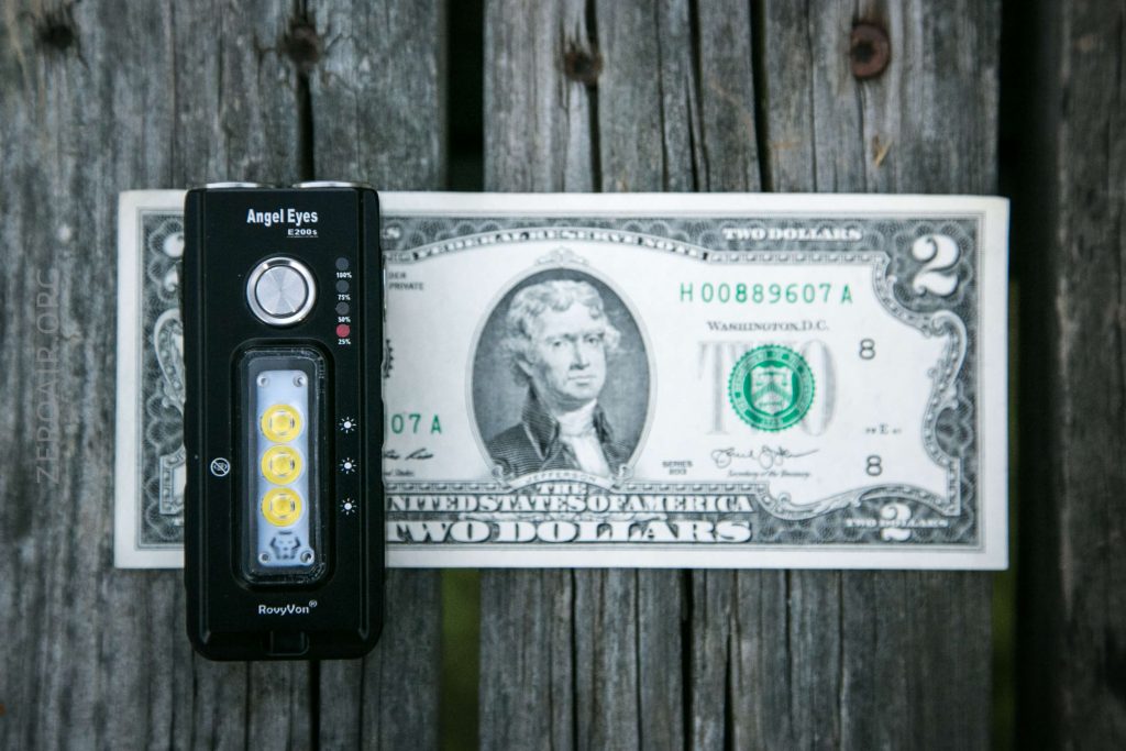 A black flashlight labeled Angel Eyes is placed on top of a two-dollar bill featuring Thomas Jefferson, which is laid on a weathered wooden surface.