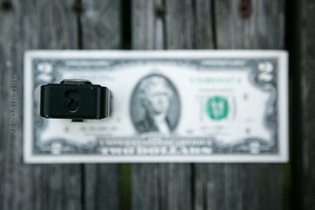 A small black rectangular object is positioned vertically in front of a two-dollar bill that is lying flat on a wooden surface. The bill and the wood are out of focus.