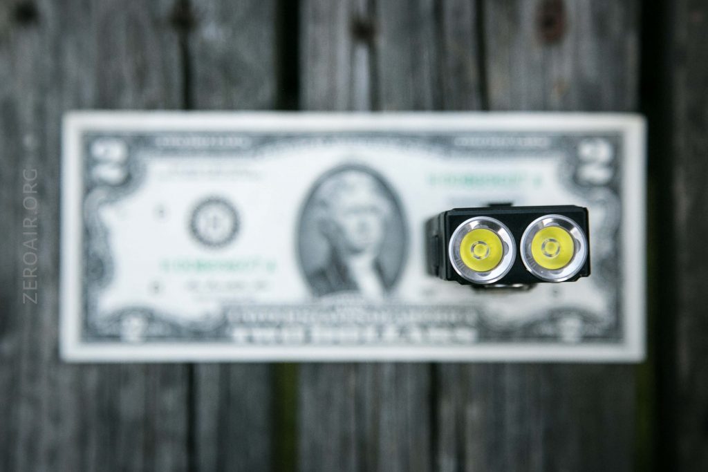 A two-dollar bill lies on a wooden surface. In the foreground, a small flashlight with two LED bulbs is positioned above the bill, mostly in focus, while the bill is blurred.