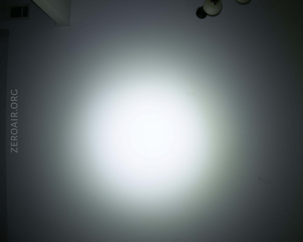 A white wall illuminated by a bright circular beam of light, with a ceiling vent in the upper left corner, a light fixture in the upper right, and the text ZEROAIR.ORG faintly visible on the left side.