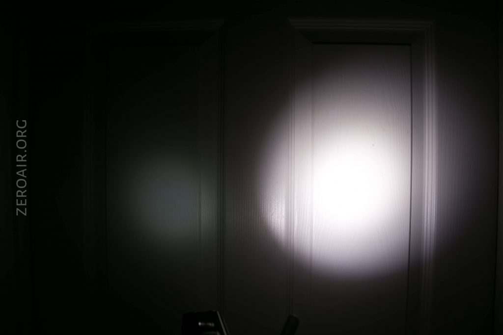 A bright flashlight beam shines on a closed white door in a dark room, creating a circular area of light in the center and a faint, smaller circle to the left. The word ZEROAIR.ORG appears vertically on the left side.