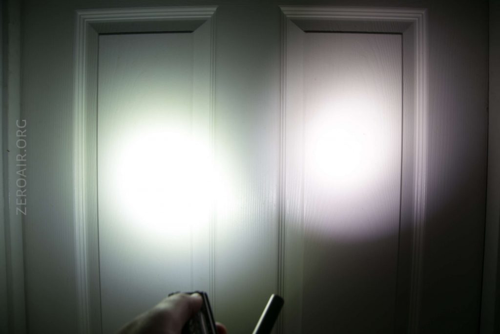 A hand holds a flashlight pointing at a white paneled door, creating two bright circular spots of light on the door’s surface. The text ZEROAIR.ORG is visible along the left side.