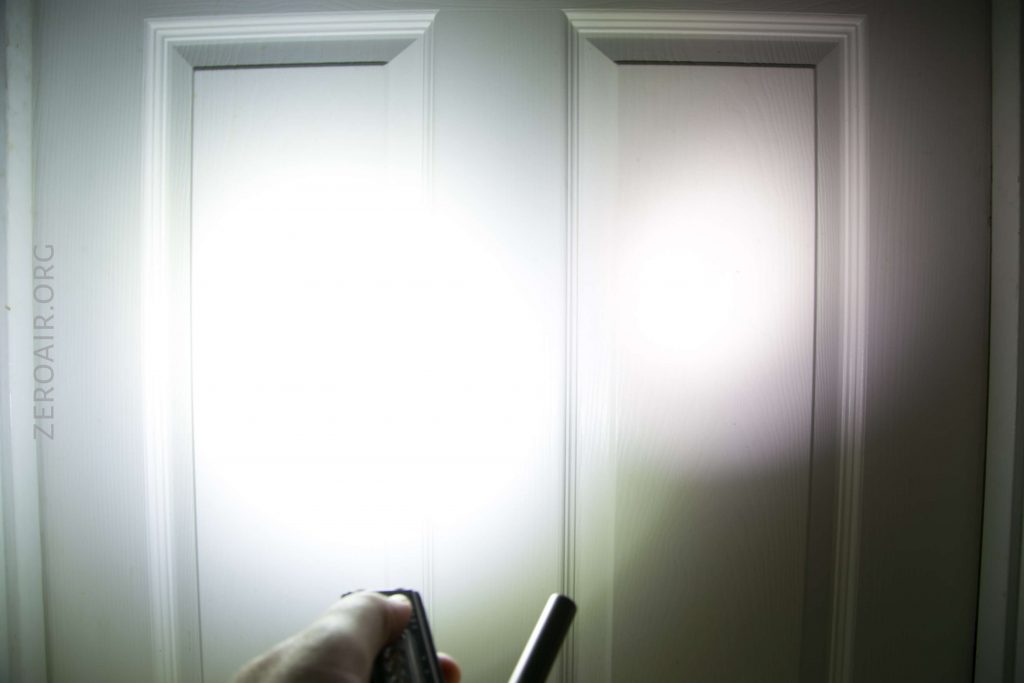 A hand holding a flashlight is pointed at a white paneled door in a dark room, creating two bright circular beams of light on the door’s surface.