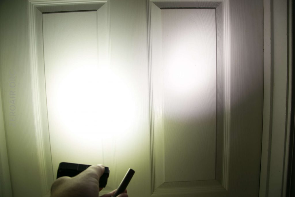 A hand holding a flashlight points a bright beam of light at a white paneled door, illuminating two panels with strong circular light reflections.