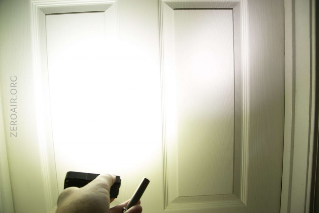 A hand holds a flashlight shining a bright beam onto a white paneled door, causing a large illuminated spot on the doors surface. The text ZEROAIR.ORG is visible on the left side of the image.
