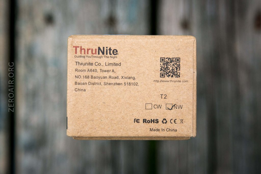 A cardboard box with the ThruNite logo, company details, a QR code, and product information printed on it, placed on a wooden surface. The box is labeled T2 and has various certification icons.