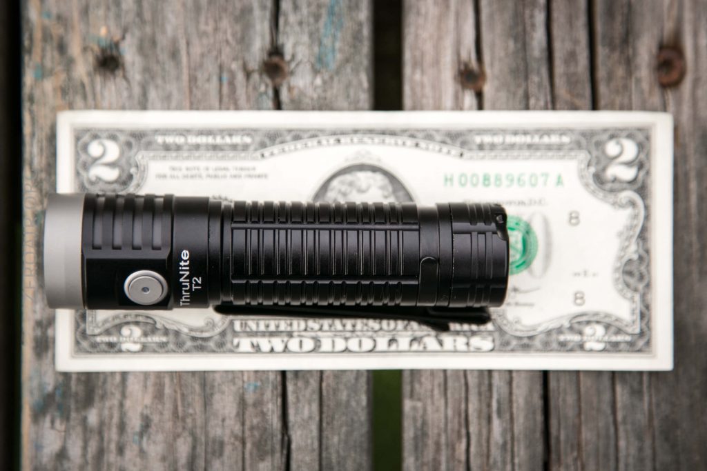 A black ThruNite TN12 flashlight is placed horizontally on top of a U.S. two-dollar bill, which rests on a weathered wooden surface.