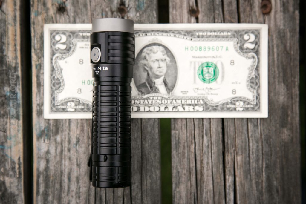 A black ThruNite T2 flashlight is placed vertically on top of a U.S. two-dollar bill, both lying on a weathered wooden surface. The flashlight covers part of the bills center.