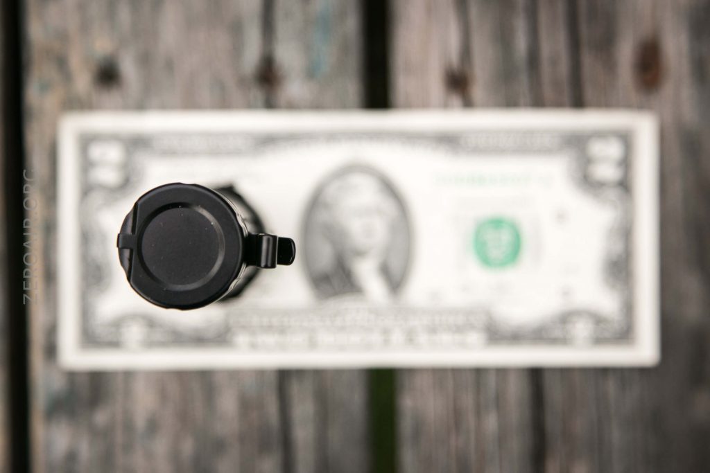 A close-up, top-down view of a black handgun pointing toward the camera, with a US dollar bill lying flat on a wooden surface in the background. The dollar bill is out of focus.