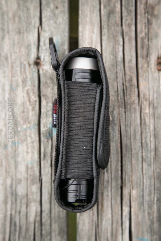 A black flashlight in a holster with a zipper, viewed from above, placed on a weathered wooden surface.