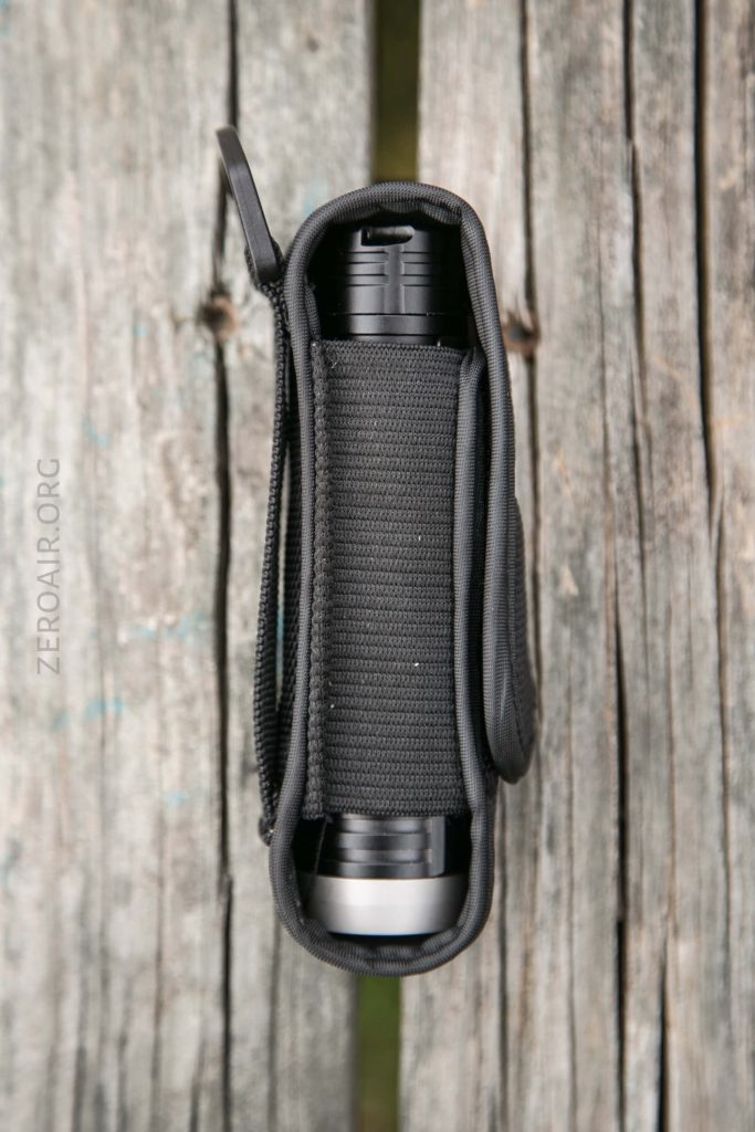 A black flashlight inside a black nylon holster, viewed from above, resting on weathered wooden planks. The holster is snug around the flashlight, with the lens partially visible at the bottom.