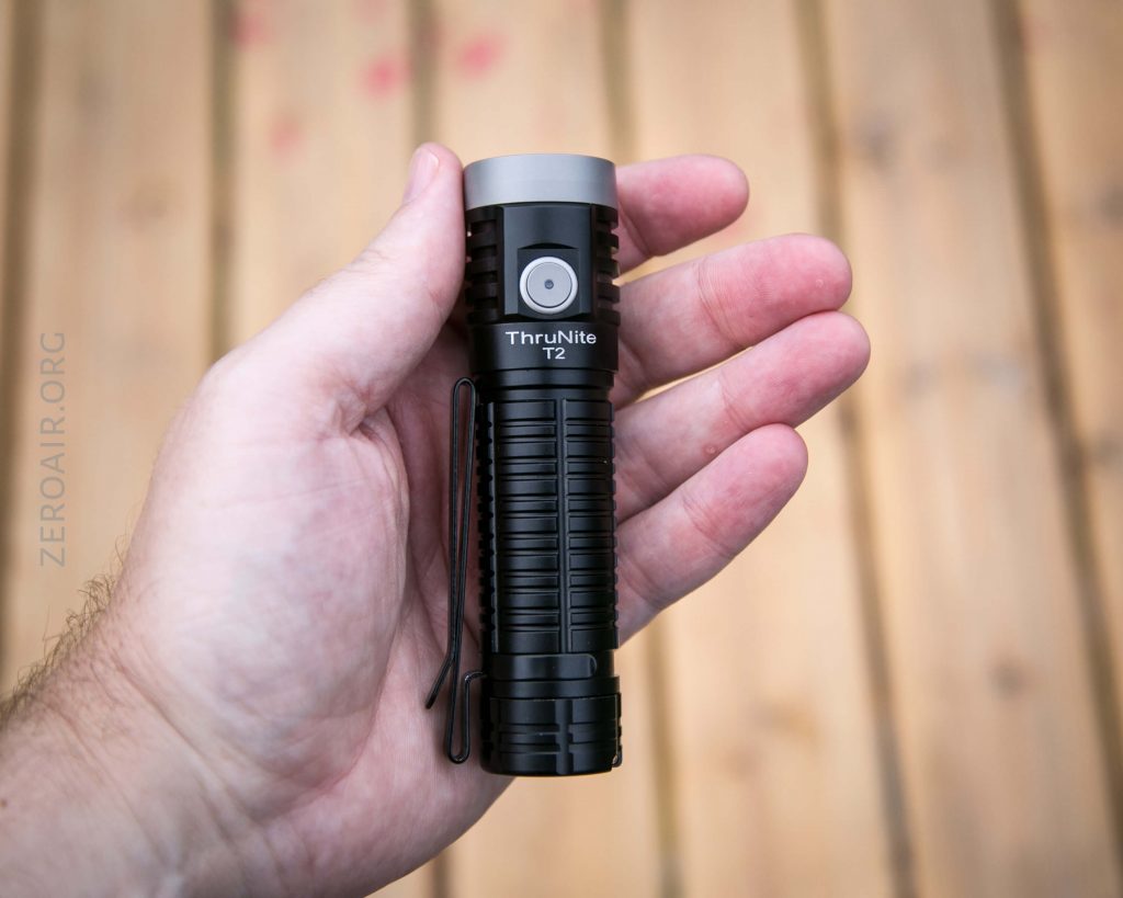 A hand holds a black ThruNite T2 flashlight with a textured grip and side button, against a blurred wooden background.