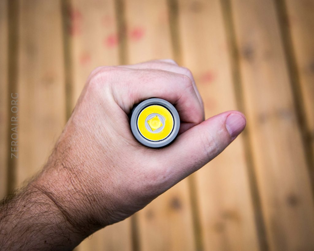 A hand is holding a flashlight viewed from above, with the lens and yellow LED facing the camera. The background is a wooden surface with parallel planks.