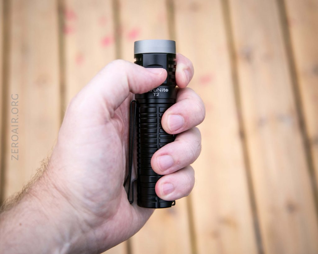 A hand holding a small black flashlight with a ridged grip and a gray top, positioned over a blurred wooden surface. The word ZERONAIR.ORG is faintly visible on the left side.
