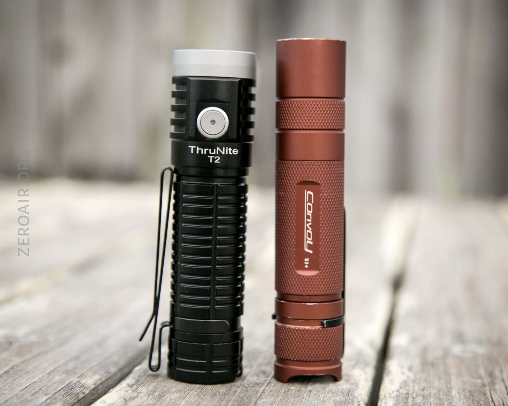 Two flashlights stand upright on a wooden surface outdoors. The left flashlight is black and labeled ThruNite T2. The right flashlight is copper-colored and labeled Sofirn. Both have textured grips and clips.