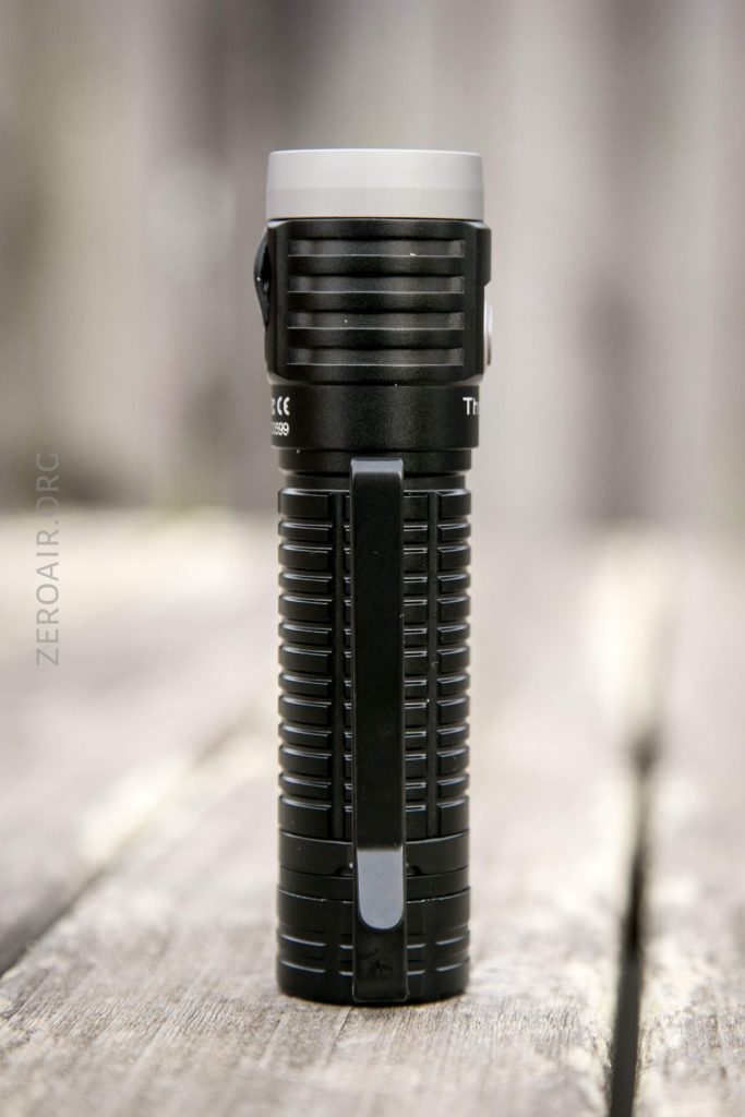 A black, cylindrical flashlight with a textured grip and a metal pocket clip stands upright on a weathered wooden surface. The background is blurred.
