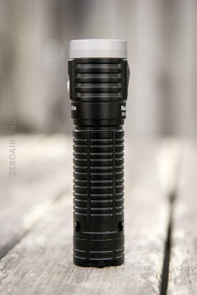 A black, cylindrical flashlight with horizontal and vertical grooves stands upright on a weathered wooden surface, with a blurred background.