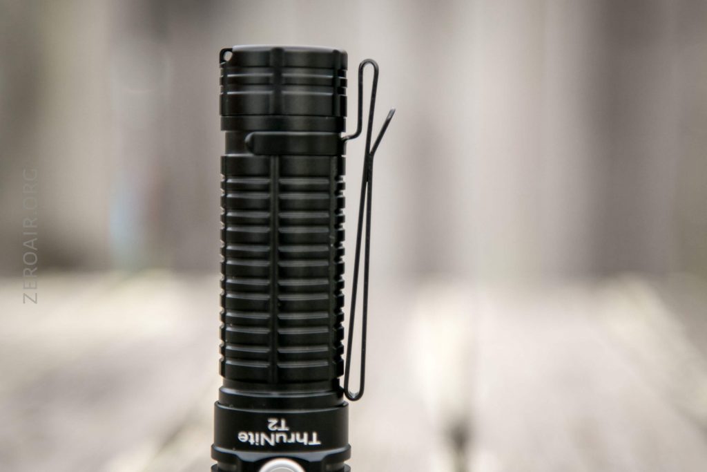 A close-up of a black ThruNite T2 flashlight standing upright on a wooden surface, shown from the side with a metal pocket clip visible. The background is blurred.