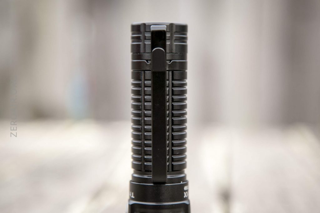 A close-up view of a black, ridged flashlight standing upright on a light wooden surface with a blurred background. A sturdy metal pocket clip is attached along the length of the flashlight.