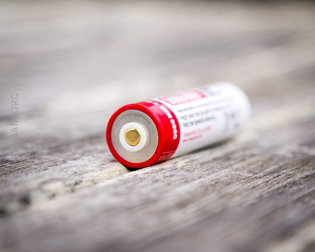 A close-up of a single AA battery lying on its side on a wooden surface, with the positive terminal facing the camera. The battery has a red and white label.