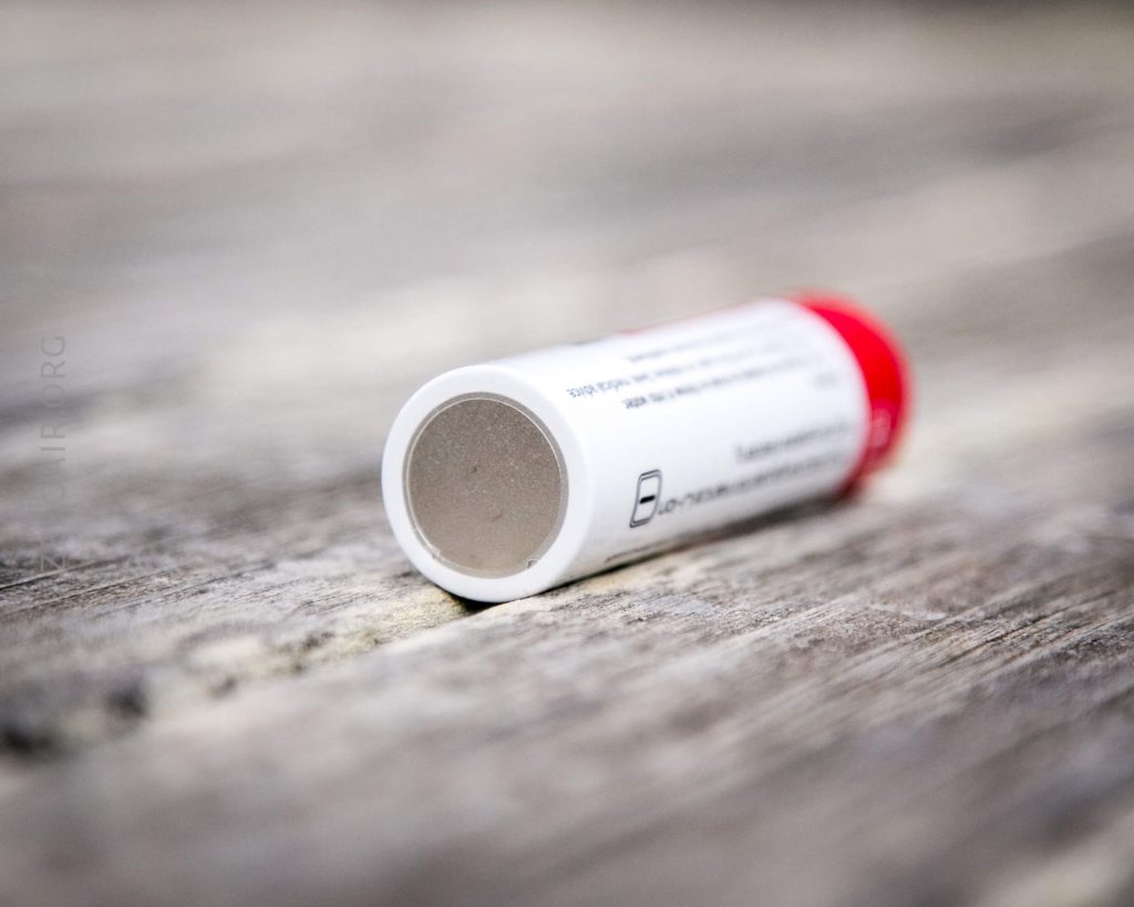A close-up of a small cylindrical container with a red cap, lying on its side on a weathered wooden surface. The label text is blurred and unreadable.