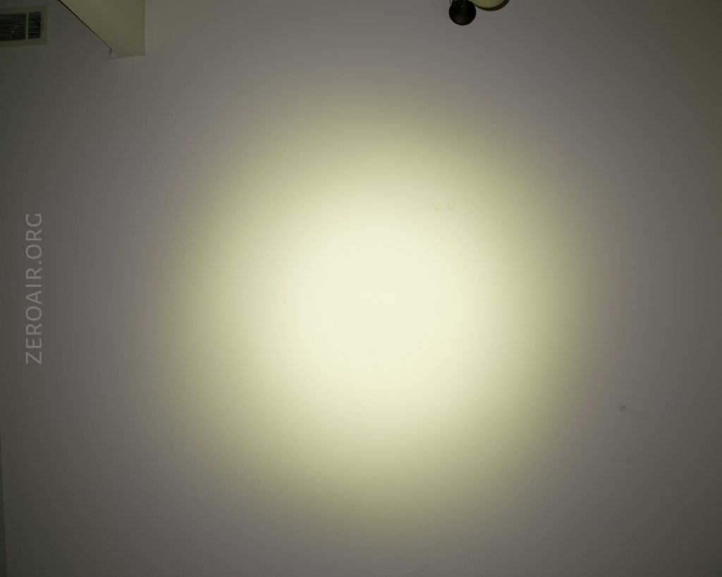 A circular beam of yellowish light is projected onto a plain white wall. The word ZEROAIR.ORG is vertically printed on the left side of the image. There is a ceiling vent and light fixture partially visible at the top.