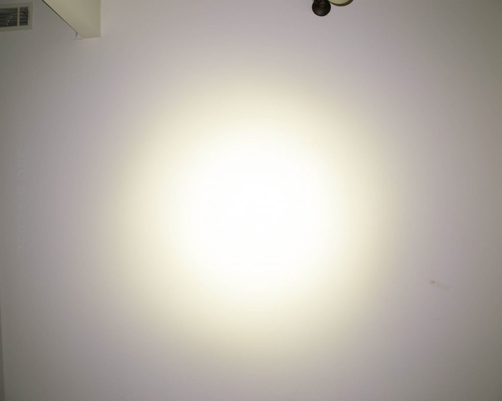 A bright white light is shining on a plain off-white wall, creating a circular glowing effect in the center. A small portion of a ceiling fixture is visible in the top right corner.