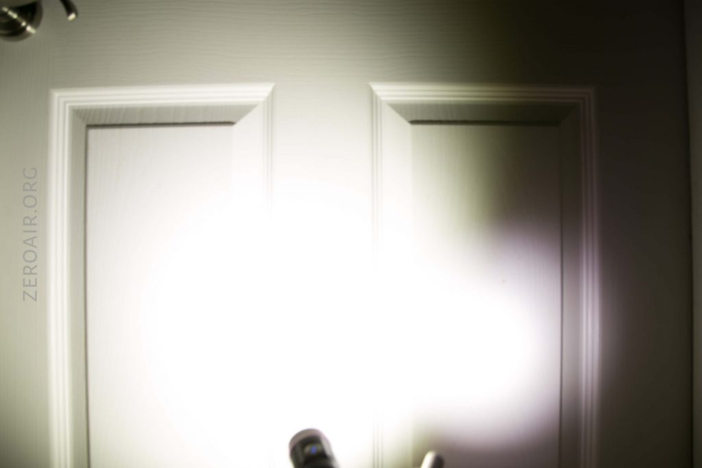 A white door with rectangular panels is illuminated by a bright flashlight beam in the center, creating a glare and shadow. The text ZEROAIR.ORG is visible vertically on the left side.