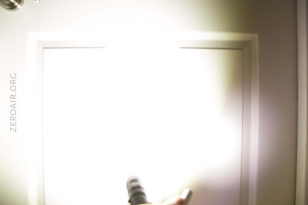 A bright white light from a flashlight shines directly at a white door, causing overexposure. The word ZEROAIR.ORG is vertically printed along the left side of the image.