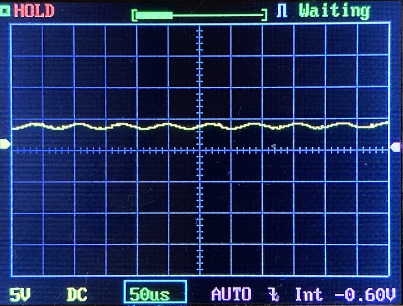 A digital oscilloscope display shows a mostly flat yellow waveform with slight fluctuations. The horizontal scale is 50 microseconds per division, and the vertical scale is 5 volts per division. The screen reads “HOLD” and “Waiting.”