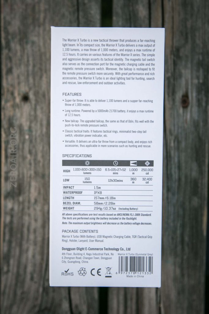 A white product box with printed specifications and features for the Warrior X Turbo tactical flashlight, including output levels, beam distance, waterproof rating, dimensions, and package contents. The box is on a wooden surface.