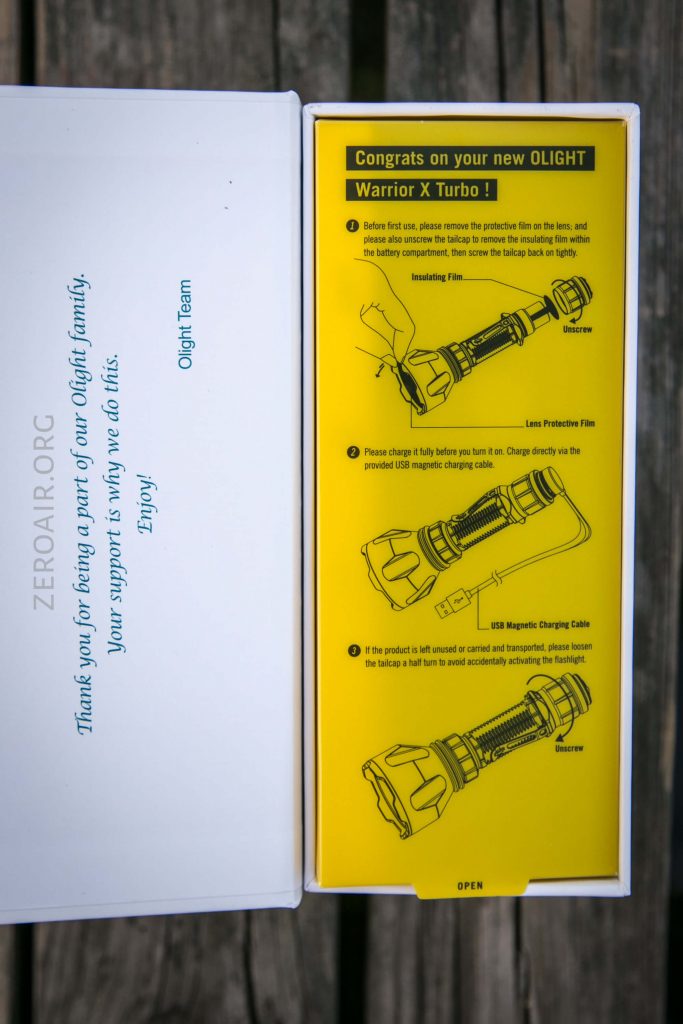 An open box containing a yellow instruction card for the OLIGHT Warrior X Turbo flashlight, showing illustrated setup steps. The left side has a printed thank-you message from the Olight Team.