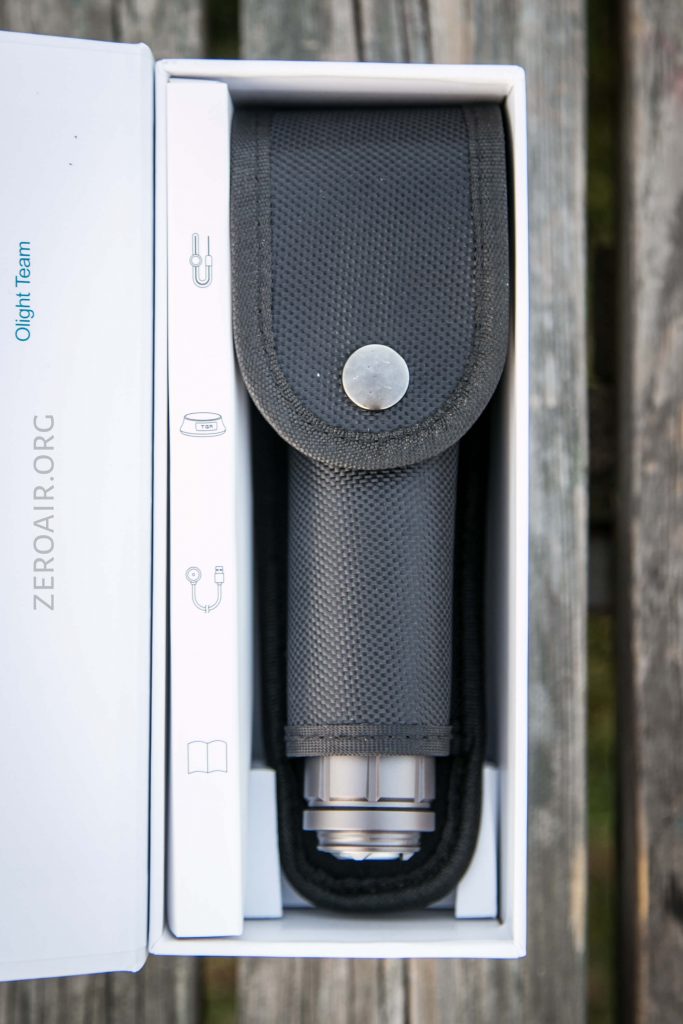 A black flashlight case with a snap button is placed in a white box. Part of a silver flashlight is visible at the bottom of the case. The box has text and simple icons printed on the inner flap.