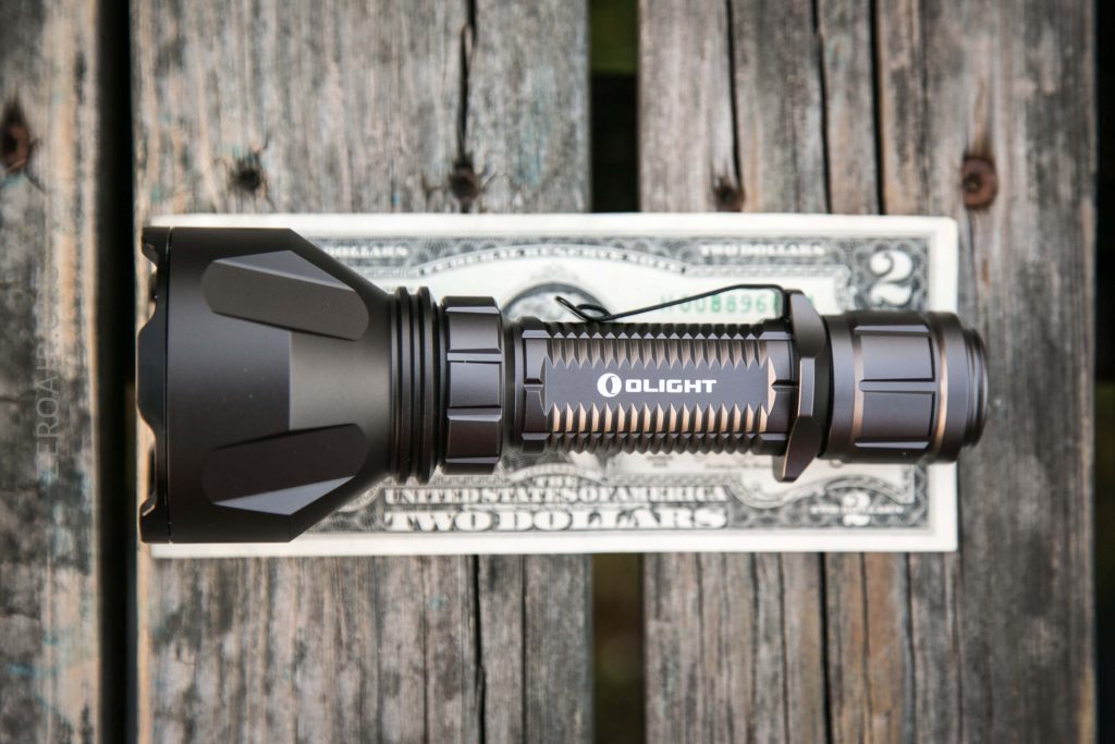 A black Olight flashlight is placed on top of a two-dollar bill on a weathered wooden surface, showing the size of the flashlight in comparison to the bill.